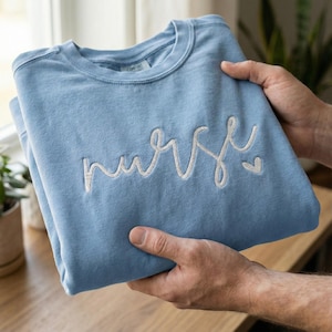 Könnte beinhalten: Hellblaues Sweatshirt mit dem Wort "nurse" in weißer Schrift auf der Vorderseite gestickt. Das Sweatshirt ist gefaltet und wird von Händen gehalten, wodurch der weiche Stoff und das Design hervorgehoben werden. Ein Geschenk für Krankenschwestern.