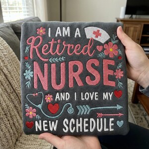 May include: A dark gray square decorative pillow with embroidered text that reads "I AM A Retired NURSE AND I LOVE MY NEW SCHEDULE." The design includes a nurse's cap, flowers, hearts, and a stethoscope.