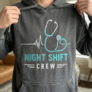 May include: Dark gray hoodie with a teal stethoscope design and the words "NIGHT SHIFT CREW" in teal. The design includes a white heartbeat line. The hoodie has a drawstring hood and a front pocket.