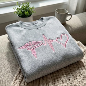 May include: A gray sweatshirt with a pink embroidered design featuring a caduceus symbol, a heartbeat line, and a heart. The sweatshirt is folded and resting on a beige knitted blanket.