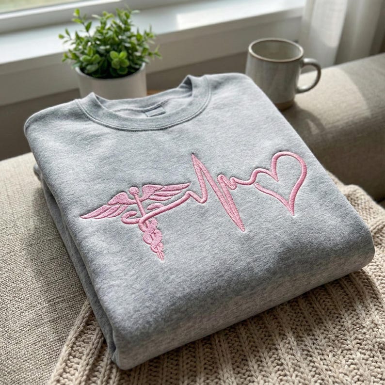 May include: A gray sweatshirt with a pink embroidered design featuring a caduceus symbol, a heartbeat line, and a heart. The sweatshirt is folded and resting on a beige knitted blanket.