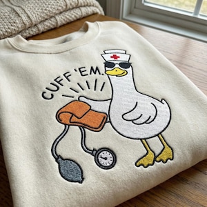 May include: Cream-colored sweatshirt with an embroidered cartoon goose wearing sunglasses and a nurse's hat. The goose is holding a blood pressure cuff. Text above the cuff reads "CUFF 'EM."