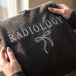 Może przedstawiać: Ciemnoszara bluza z kapturem z białym haftem "RADIOLOGY" i białym wzorem kokardy. Bluza jest złożona, ukazując szczegóły haftu. Materiał wydaje się miękki i wygodny.