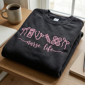 May include: A dark gray sweatshirt with the words "nurse life" in pink script, along with pink embroidered medical symbols. A coffee mug and notepad are visible in the background.