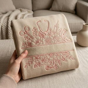 May include: A folded beige sweatshirt featuring intricate pink embroidery of medical instruments including a stethoscope, scissors, a syringe, and a thermometer, all intertwined with floral motifs and leaves, centered around a blank horizontal banner.