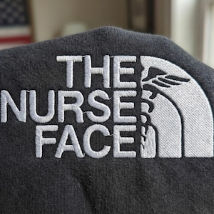 Könnte beinhalten: Nahaufnahme eines dunkelgrauen Stoffes mit weiß gesticktem Text "THE NURSE FACE". Das Design beinhaltet ein stilisiertes Berg- und Baumlogo, eine Parodie einer bekannten Marke.