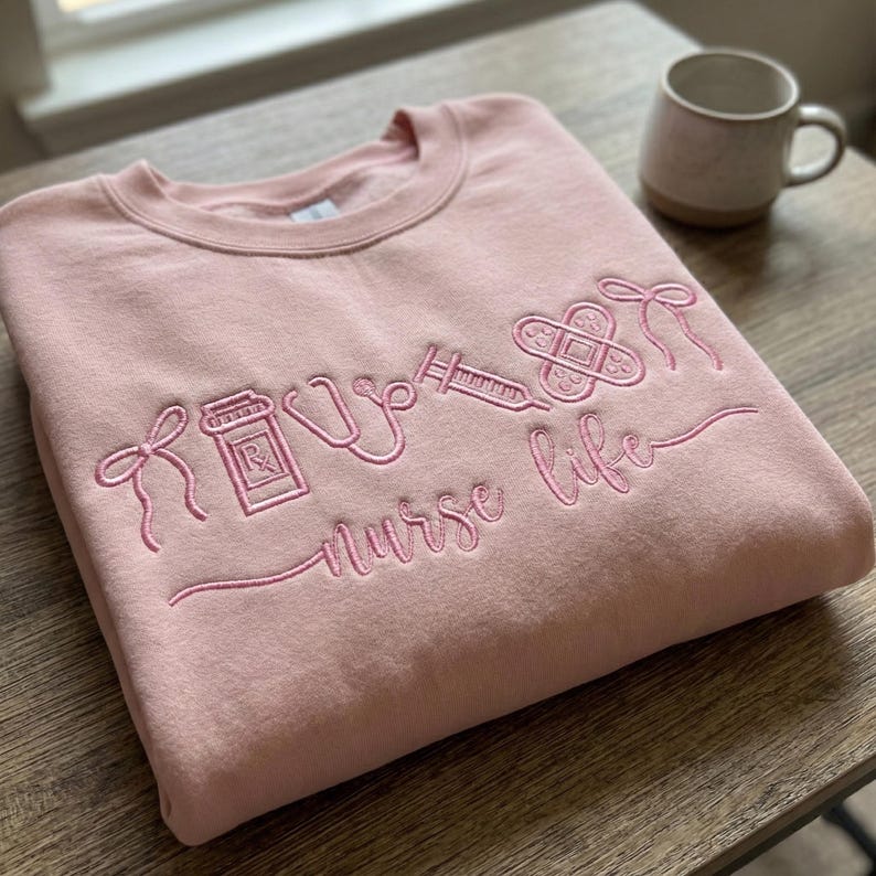 May include: A light pink sweatshirt embroidered with the words "nurse life" and medical symbols, including a stethoscope, syringe, and bandage. A small, beige mug sits in the background. The sweatshirt is folded on a wooden surface.
