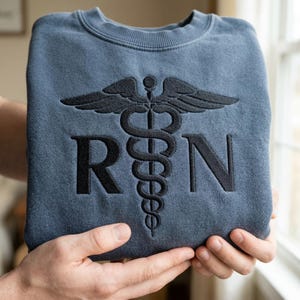 May include: A blue sweatshirt featuring a black embroidered caduceus symbol and the letters "RN". The sweatshirt is held up by a person. The design is centered on the front of the garment.