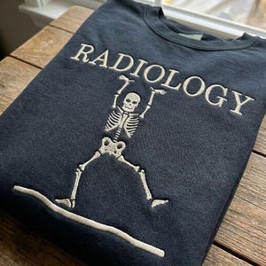 May include: Dark navy blue sweatshirt featuring the word "RADIOLOGY" embroidered in white above a white skeleton graphic. The skeleton has its arms raised. The sweatshirt is made of a soft material.