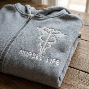 May include: A gray zip-up hoodie with a white embroidered medical symbol and the text "NURSEE LIFE". The hoodie is folded and appears to be made of a soft, comfortable material. The zipper is visible, and the hoodie is resting on a wooden surface.