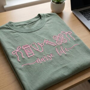 May include: Sage green t-shirt with pink embroidered medical-themed graphics and the words "nurse life." The design includes a pill bottle, stethoscope, syringe, and bandage. The shirt is folded neatly on a wooden surface.
