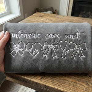 May include: A gray sweatshirt with white embroidered text and designs. The text reads "intensive care unit" above a series of embroidered images including bows, a heart with a heartbeat line, a stethoscope, and more bows.