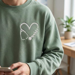 Puede incluir: Sudadera verde salvia con un diseño de corazón bordado en blanco. El corazón incorpora un estetoscopio y la palabra "nurse" en escritura cursiva. La sudadera es de cuello redondo.
