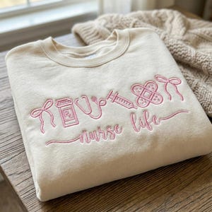 May include: Cream-colored sweatshirt with pink embroidered medical-themed graphics, including a prescription bottle, stethoscope, syringe, bandage, and bow. The words "Nurse Life" are also embroidered in pink. The sweatshirt is folded on a wooden surface.