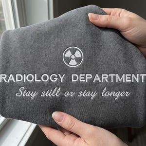 May include: A gray sweatshirt with white embroidered text and a radiation symbol. The text reads "RADIOLOGY DEPARTMENT" above the phrase "Stay still or stay longer."