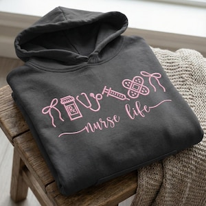 May include: Dark gray hoodie with a pink outline design featuring medical symbols and the words "nurse life." The design includes a prescription bottle, stethoscope, syringe, bandage, and a bow. The hoodie has a hood and a drawstring.