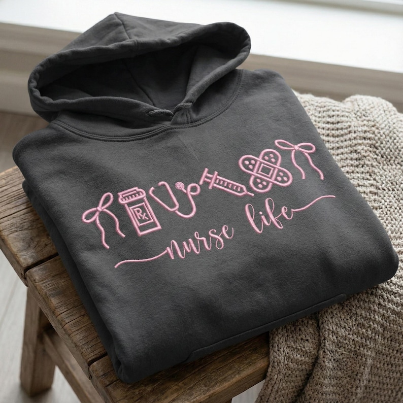 May include: Dark gray hoodie with a pink outline design featuring medical symbols and the words "nurse life." The design includes a prescription bottle, stethoscope, syringe, bandage, and a bow. The hoodie has a hood and a drawstring.