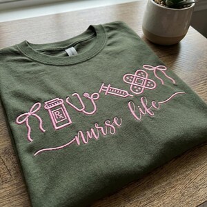 May include: Olive green t-shirt with pink embroidered medical symbols and the words "nurse life". The design includes a prescription bottle, stethoscope, syringe, and bandage. The shirt is made of a soft material.