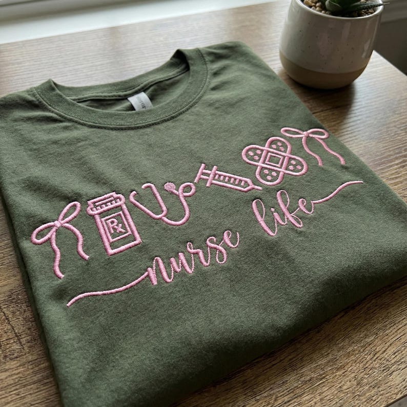 May include: Olive green t-shirt with pink embroidered medical symbols and the words "nurse life". The design includes a prescription bottle, stethoscope, syringe, and bandage. The shirt is made of a soft material.