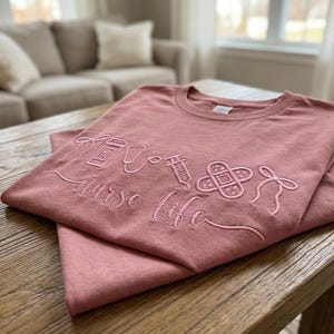May include: A dusty rose-colored t-shirt with embroidered medical-themed designs and the words "nurse life." The design includes a pill bottle, stethoscope, syringe, bandage, and a medical ribbon. The shirt is folded on a wooden table.