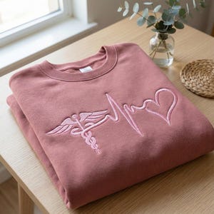 May include: A dusty rose sweatshirt with a crew neck. The front features a light pink embroidered design of a caduceus symbol, a heartbeat line, and a heart. The sweatshirt is folded neatly on a wooden surface.