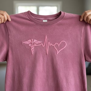 May include: A dusty rose-colored t-shirt with a pink embroidered design. The design features a caduceus symbol, a heartbeat line, and a heart shape. The shirt has a crew neck and short sleeves.