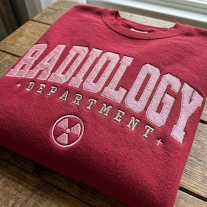 Könnte beinhalten: Ein weinrotes Sweatshirt mit dem Wort "RADIOLOGY" in großen, hellrosa gestickten Buchstaben auf der Brust. Darunter sind die Worte "DEPARTMENT" in kleinerer, grauer Schrift gestickt. Darunter ist ein Strahlungssymbol gestickt.