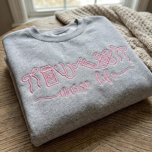 May include: A gray sweatshirt with pink embroidered medical symbols and the words "nurse life". The design includes a pill bottle, stethoscope, syringe, and bandage. The sweatshirt is folded on a wooden surface.