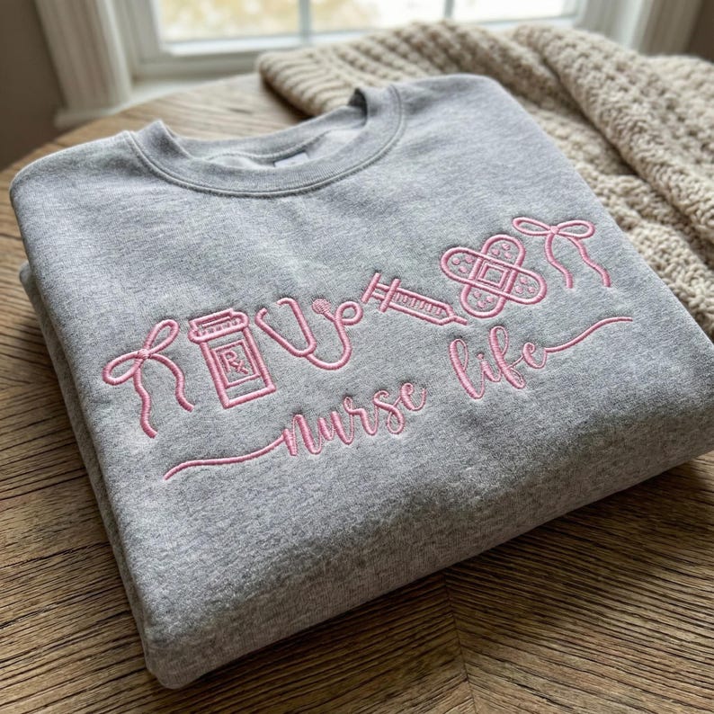 May include: A gray sweatshirt with pink embroidered medical symbols and the words "nurse life". The design includes a pill bottle, stethoscope, syringe, and bandage. The sweatshirt is folded on a wooden surface.