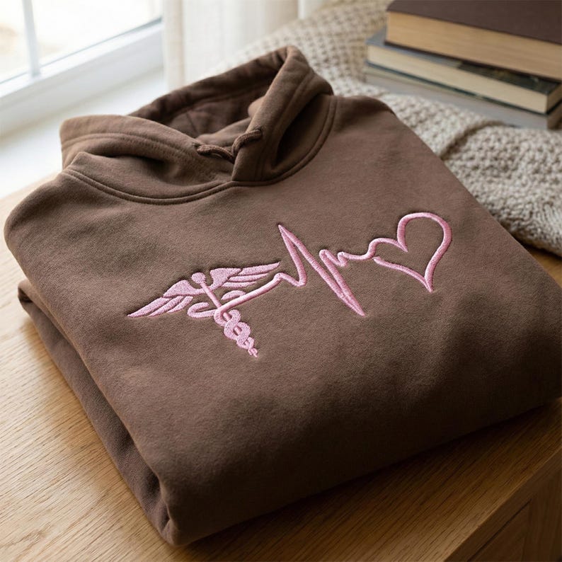 May include: A brown hoodie with a pink embroidered medical symbol and heartbeat design. The hoodie is folded neatly, showcasing the detailed stitching of the emblem. The soft fabric and color suggest comfort and a medical theme.
