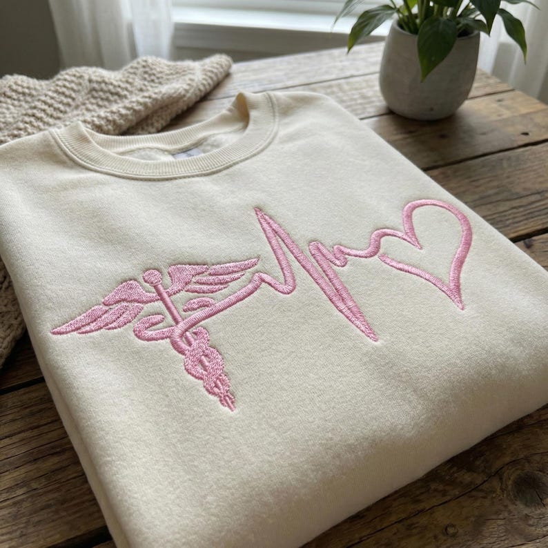 May include: Cream-colored sweatshirt with a pink embroidered design. The design features a medical caduceus symbol, a heartbeat line, and a heart shape. The sweatshirt is folded on a wooden surface.