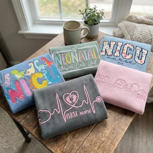 May include: A collection of embroidered sweatshirts in various colors, including blue, gray, and pink. Each sweatshirt features text and designs related to neonatal intensive care, such as "NICU nurse" and "Neonatal Intensive Care Unit."
