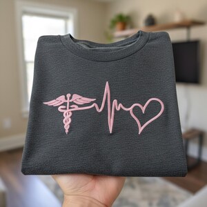 May include: Dark gray sweatshirt featuring a pink embroidered design of a medical symbol, a heartbeat line, and a heart. The sweatshirt is folded, showcasing the design prominently. The fabric has a textured appearance.