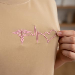 May include: A tan-colored sweatshirt with a pink embroidered design. The design features a medical symbol, a heartbeat line, and a heart shape. The sweatshirt is made of a soft material.