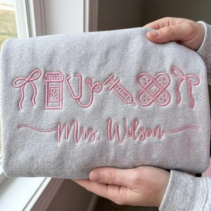 May include: A light gray sweatshirt with pink embroidered medical symbols, including a pill bottle, stethoscope, syringe, and bandage. The name "Mrs. Wilson" is also embroidered in pink. The sweatshirt is held by a person.
