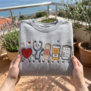 May include: A heather gray crewneck sweatshirt featuring an embroidered design of five anthropomorphic medical icons holding hands: a red heart, a blue stethoscope, a tan adhesive bandage, an orange pill bottle, and a green clipboard with a pencil.
