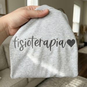 May include: A light gray heathered sweatshirt featuring the word fisioterapia embroidered in dark gray cursive script, followed by a small, solid dark gray embroidered heart symbol on the right side of the text.
