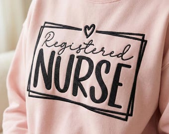 Puede incluir: Sudadera rosa claro con la frase "Registered Nurse" en negro, dentro de un rectángulo negro, y un pequeño corazón encima de la palabra "Registered". La sudadera tiene mangas largas y cuello redondo.