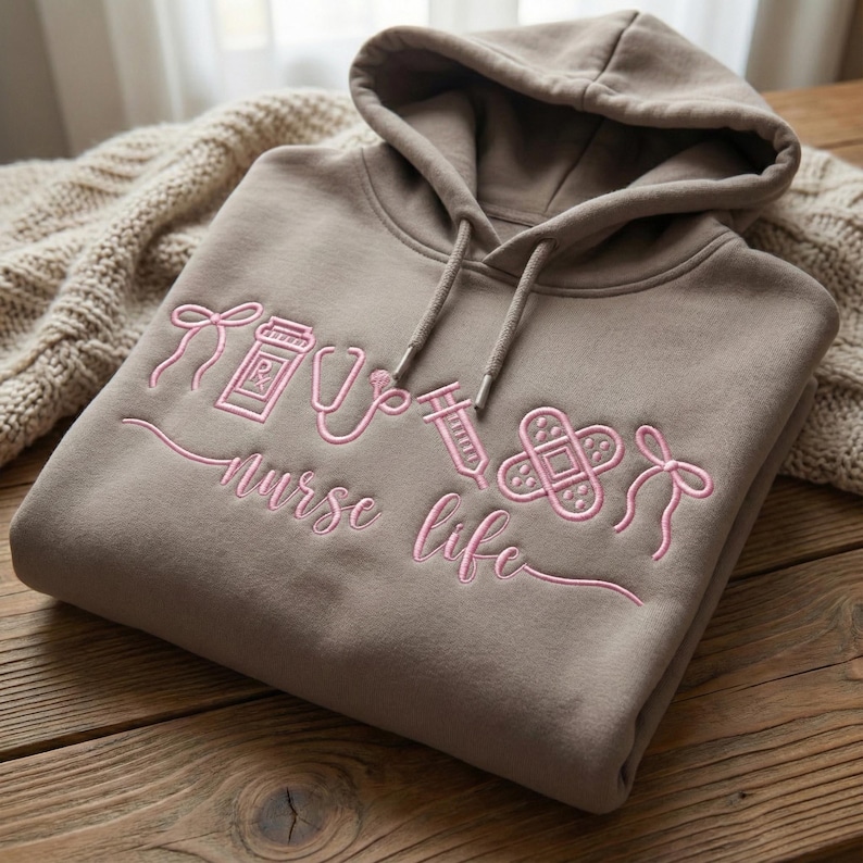 May include: A taupe hoodie with a drawstring hood. The front features pink embroidered medical symbols and the words "nurse life". The design includes a prescription bottle, stethoscope, syringe, bandage, and bow.