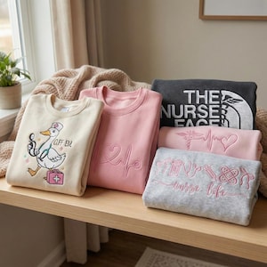 May include: Several folded sweatshirts in beige, pink, and grey. The designs include a goose in a nurse's hat, a heartbeat, and the words "Nurse Life". The sweatshirts are stacked on a wooden surface.
