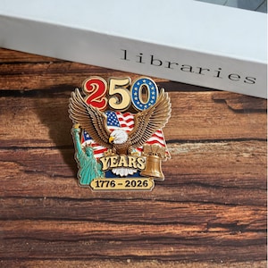 K&ouml;nnte beinhalten: Eine Gedenkanstecknadel mit einem Wei&szlig;kopfseeadler mit ausgebreiteten Fl&uuml;geln, der amerikanischen Flagge und der Freiheitsstatue. Auf der Anstecknadel steht "250 YEARS 1776-2026". Die Anstecknadel befindet sich vor einem Holzgrund.