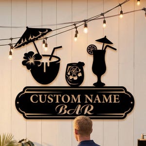 May include: Black metal sign with the text "CUSTOM NAME BAR" and cocktail illustrations. The sign is mounted on a white wall, with string lights above. A person stands near a wooden bar table.