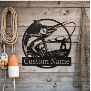 May include: A black metal wall sign featuring a circular frame with a marlin fish jumping out of the water and a silhouette of a person fishing. Below the circular design is a rectangular plaque with the text Custom Name in serif font.