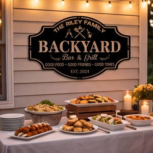 Puede incluir: Un letrero de metal negro con un borde decorativo que presenta el texto THE RILEY FAMILY BACKYARD BAR & GRILL, GOOD FOOD GOOD FRIENDS GOOD TIMES y EST. 2024, junto con iconos de una espátula, un tenedor y bebidas, montado en una pared clara.