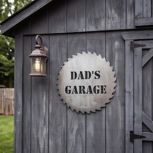Könnte beinhalten: Ein kreisförmiges Metallschild in Form eines Sägeblatts mit der schwarzen Aufschrift DAD'S GARAGE, montiert an einer Wand aus dunkelgrauen vertikalen Holzplanken neben einer bronzefarbenen Außenwandleuchte mit einem Glasschirm.