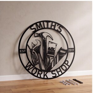 May include: A circular black metal wall sign featuring the text SMITH'S WORK SHOP and EST 2017. The center design displays a hammer, wrench, paintbrush, and hand saw. Included are several small metal bolts, spacers, and a black mounting bracket.