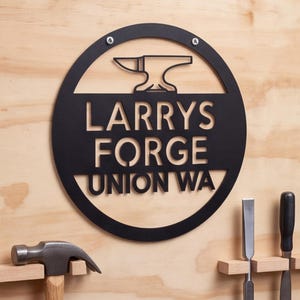 May include: A circular black metal sign featuring a cutout anvil icon at the top and the text LARRYS FORGE UNION WA in the center. The sign is mounted with two silver screws and displays a clean, industrial design with bold lettering.