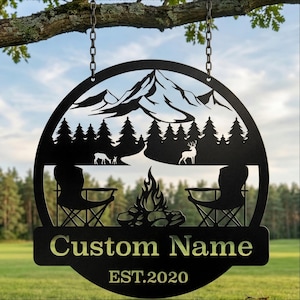 Può includere: Cartello da campeggio in metallo nero con una scena di montagna e foresta, due sedie e un falò. Il cartello include il testo "Custom Name" e "EST.2020". È appeso a un ramo.