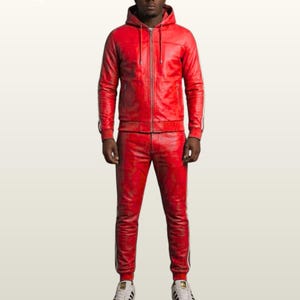 May include: A red leather tracksuit featuring a hooded zip-up jacket and matching trousers. Both pieces have white side stripes. The jacket includes a front zipper and drawstring hood. The trousers have a fitted cut. White sneakers complete the outfit.