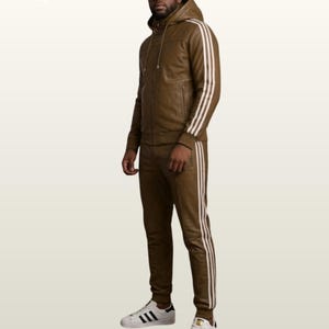 May include: A brown leather tracksuit featuring a hooded zip-up jacket and matching trousers. Both pieces are accented with three white vertical stripes along the sleeves and legs. The outfit is paired with white trainers featuring black stripes.